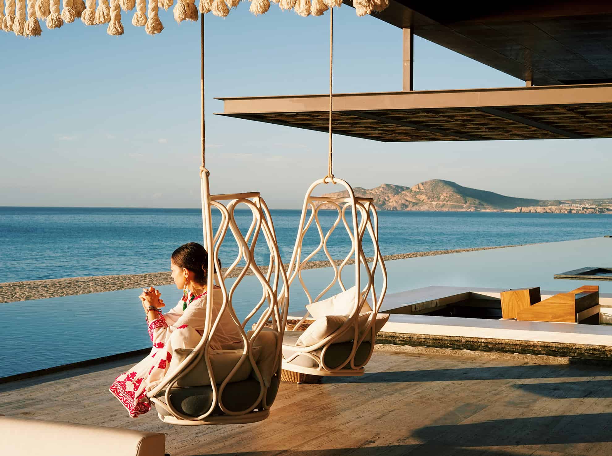 Woman in white and pink dress, hands clasped, sitting in hanging chair, looking out to the ocean