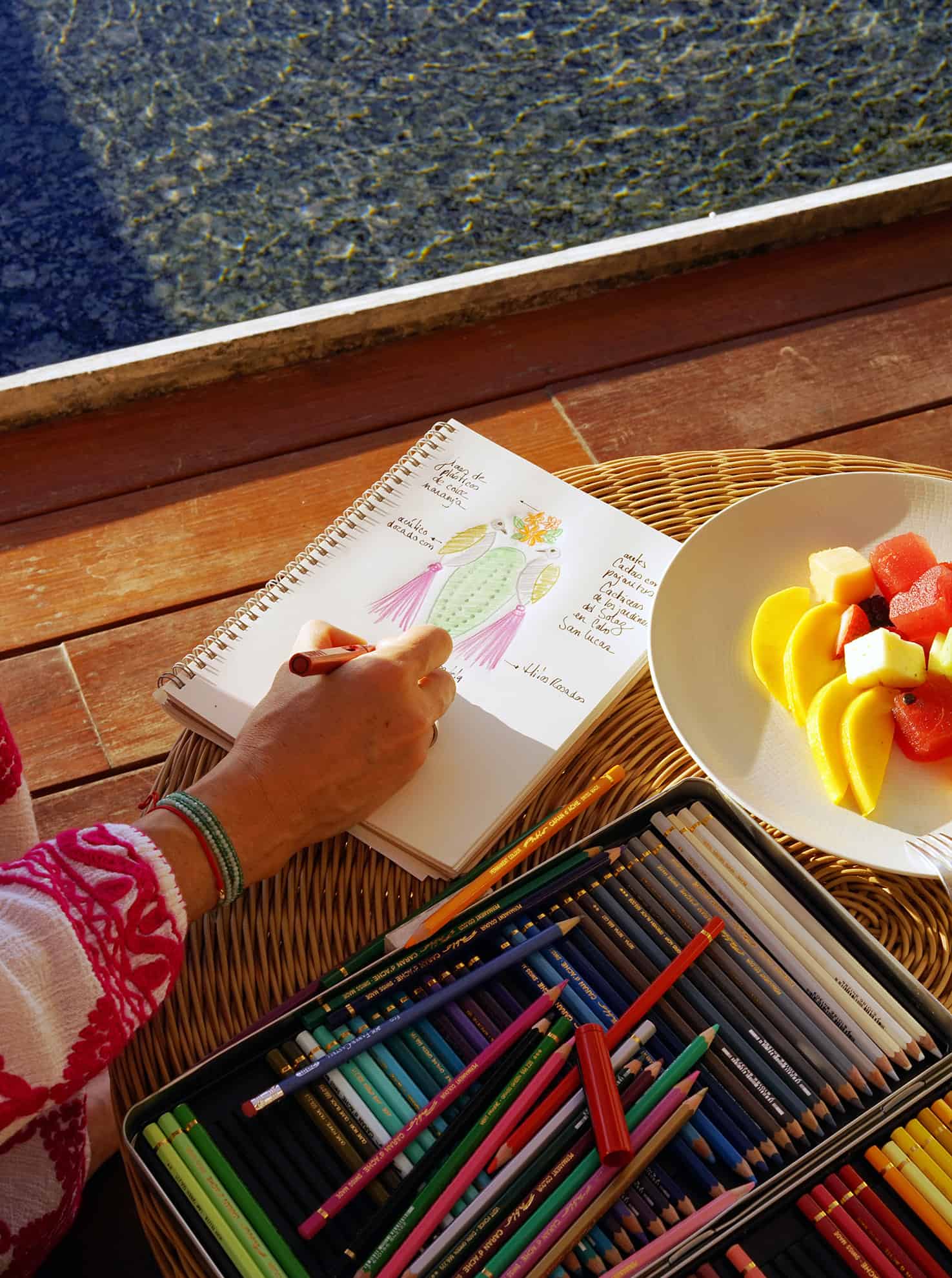 Woman's hand drawing in a notebook next to a box of colored pencils and a plate of fresh fruit