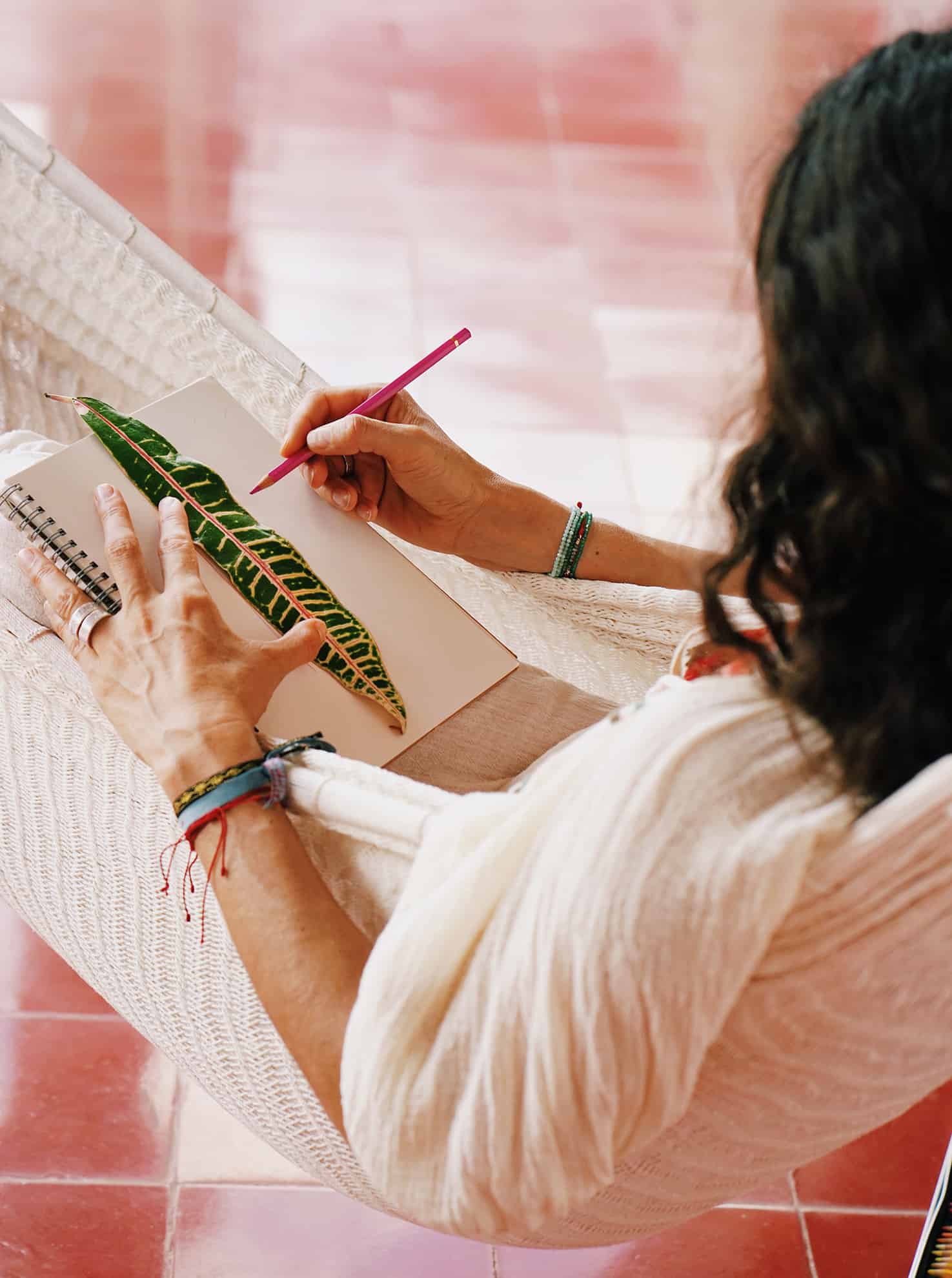 Side view of woman in white lying in white hammock tracing a tropical leaf on a notebook