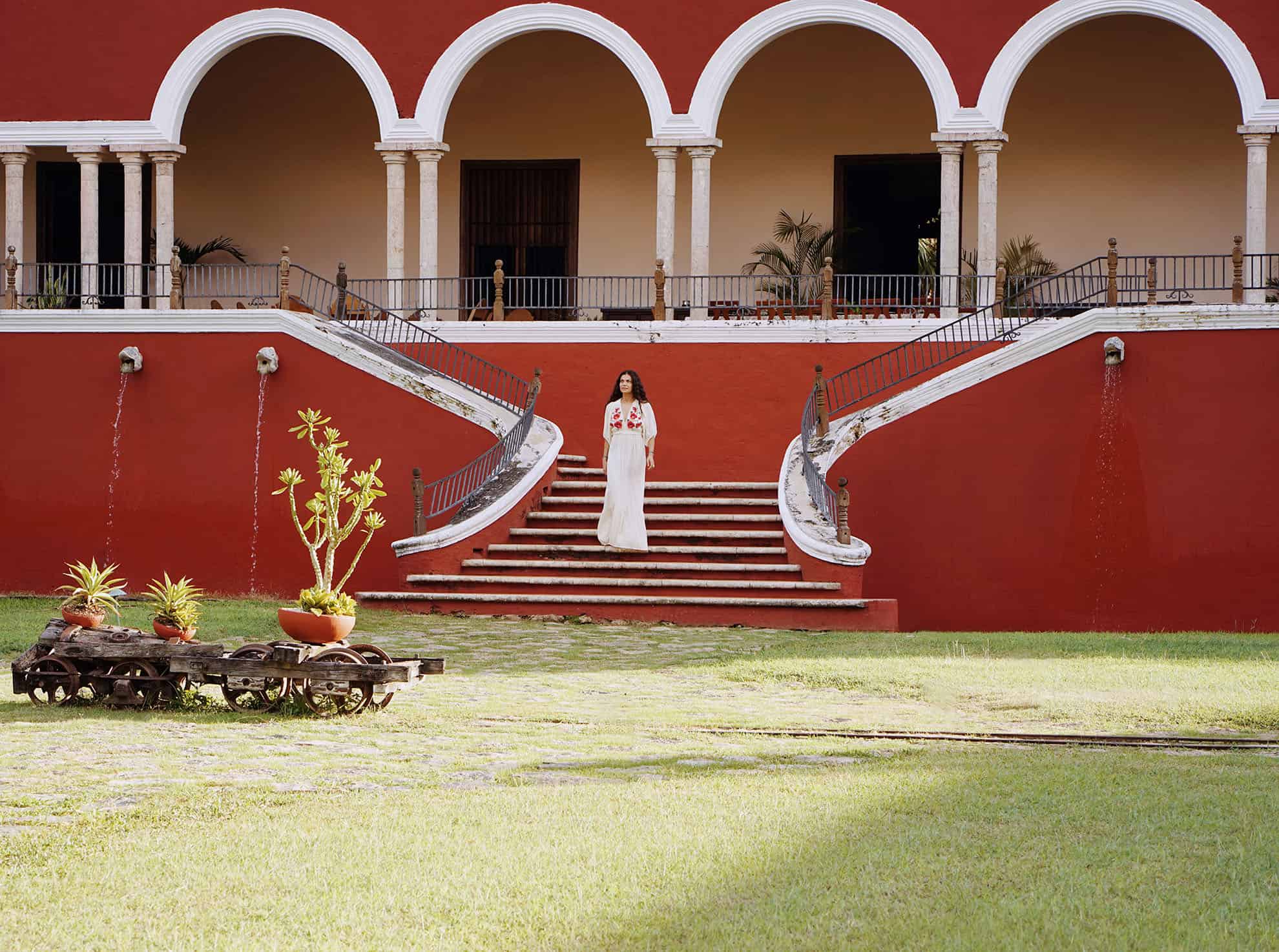 Woman in long white dress standing on steps of red building with large white archways