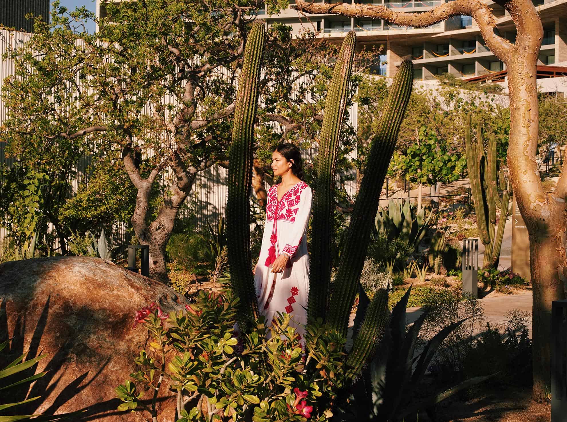 Woman in white & red traditional dress walking through a garden area with cacti, trees and rocks