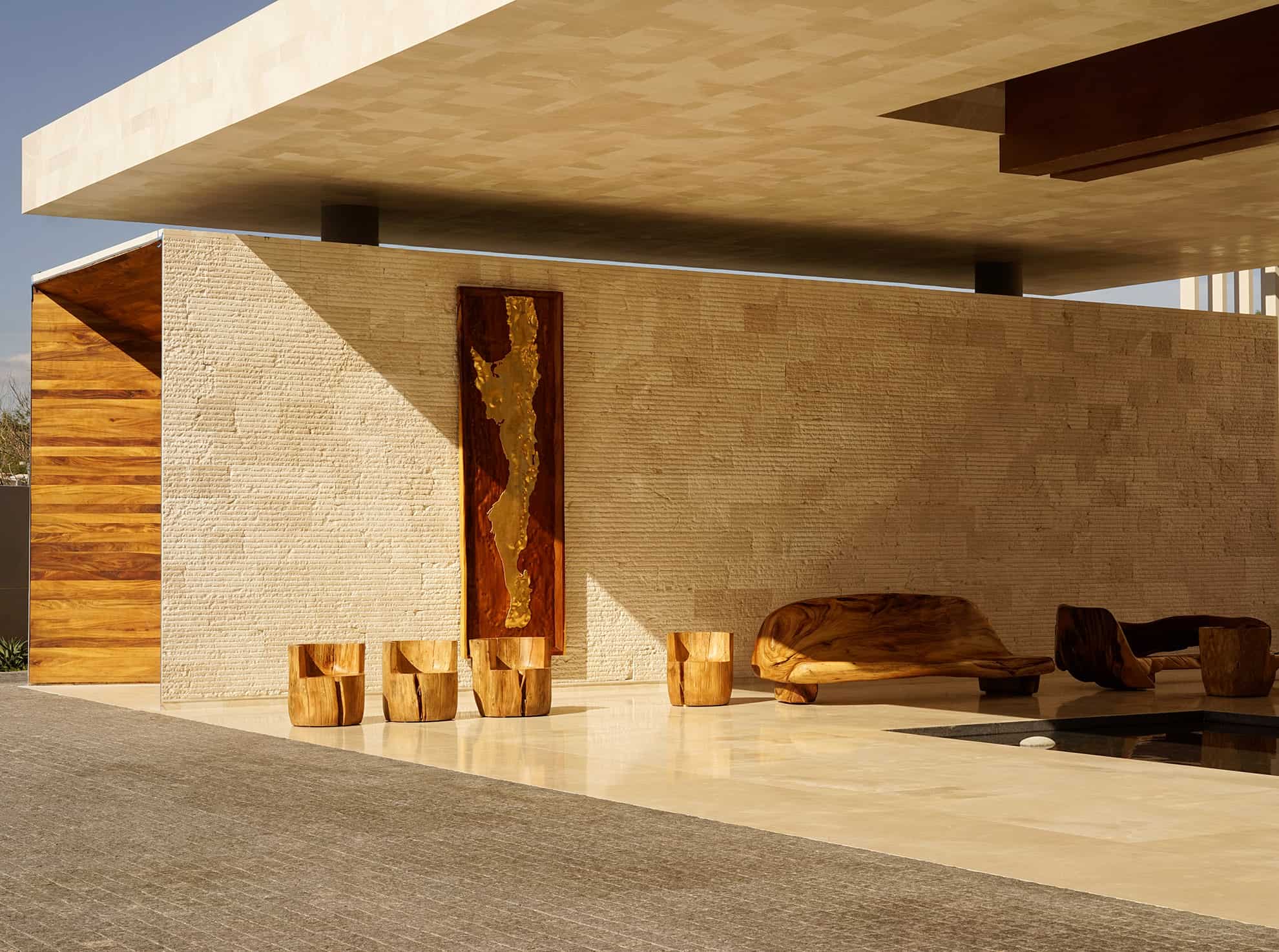 Gold and brown art piece on wall behind wood carved bench & stools next to a small pool