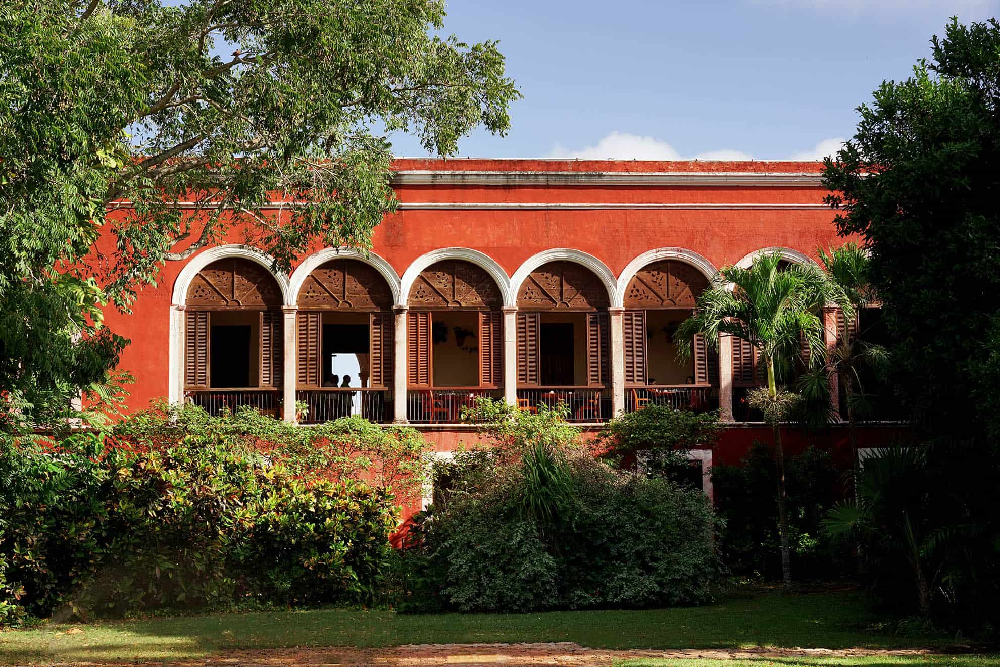Red building with shuttered arched windows on 2nd floor lined with tall trees and foliage