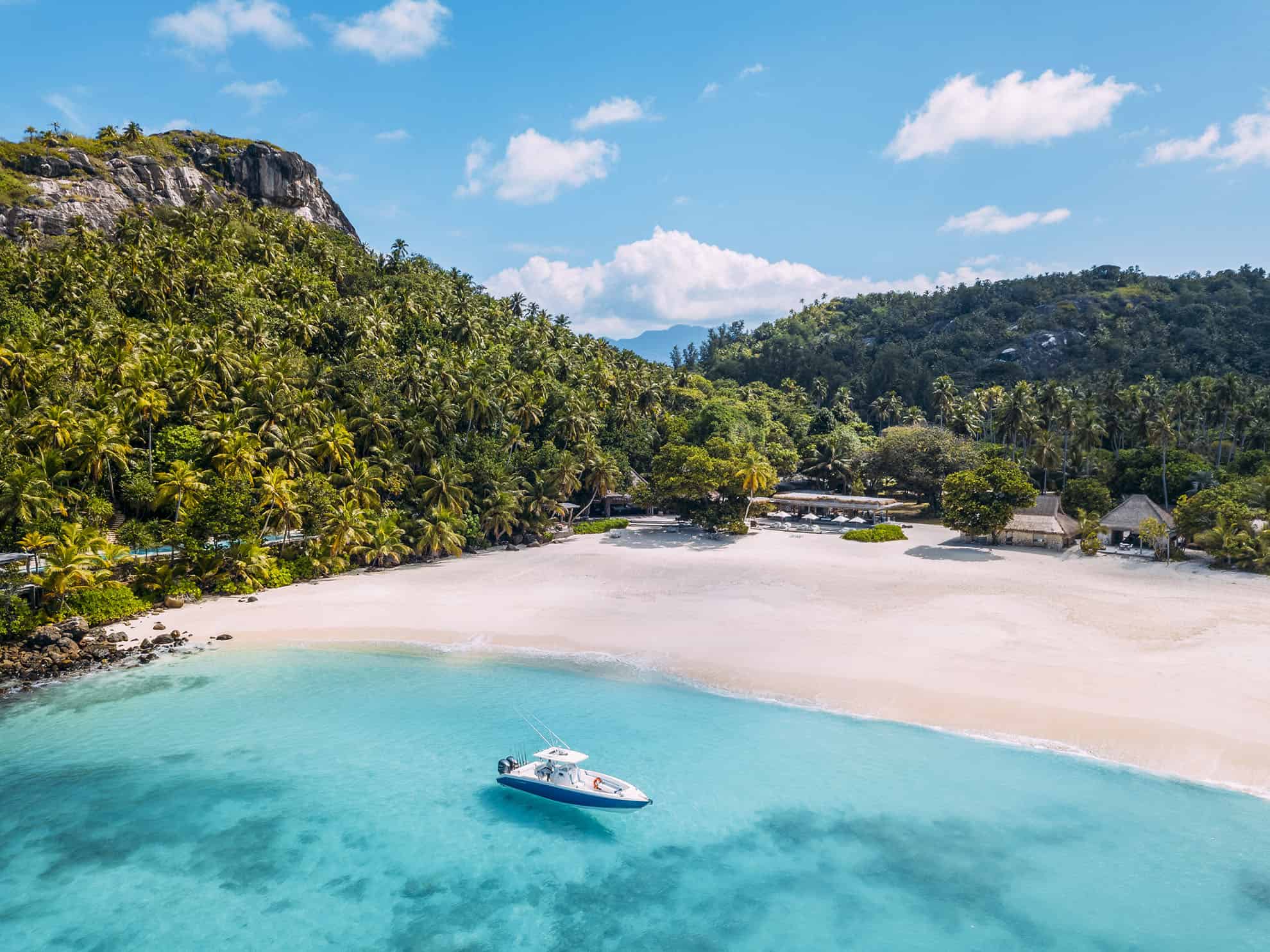 Aerial view of powerboat on clear blue ocean next to white sand beach along tree-covered cliffs