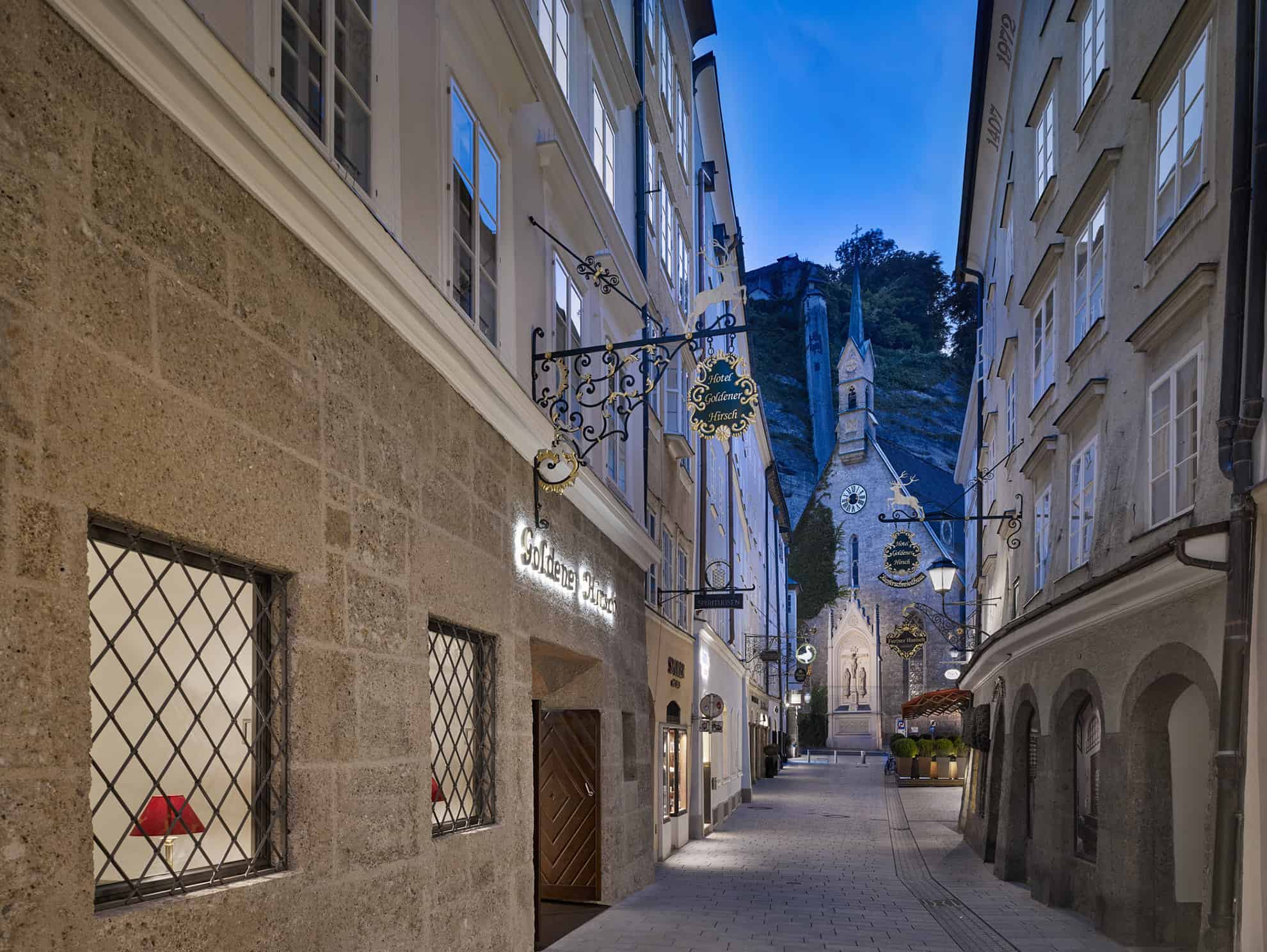 Narrow alley with dangling Hotel Goldener Hirsch sign; Churchfront with steeple at end