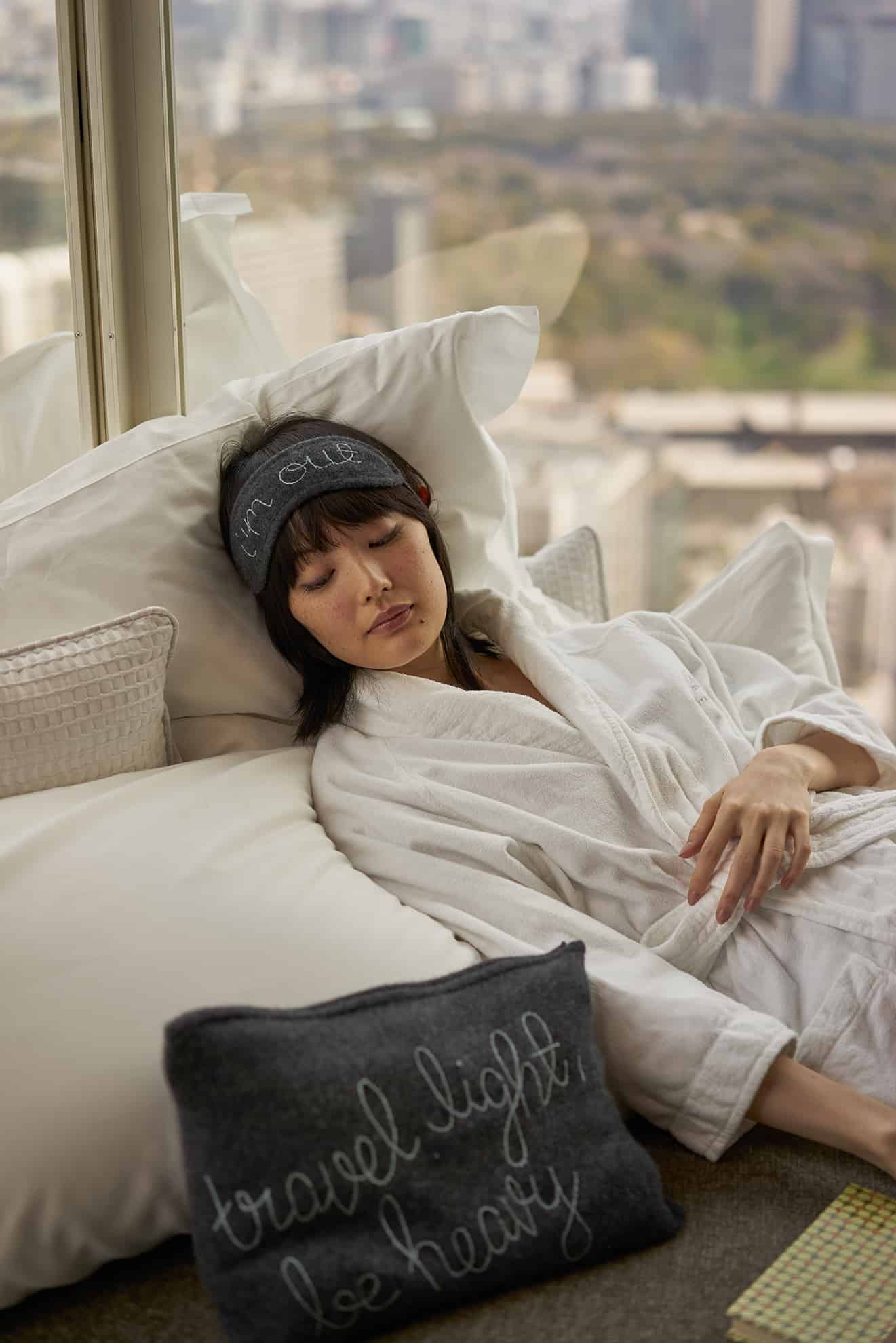 Woman in white robe napping on a pile of pillows next to a window wearing a sleep mask on forehead