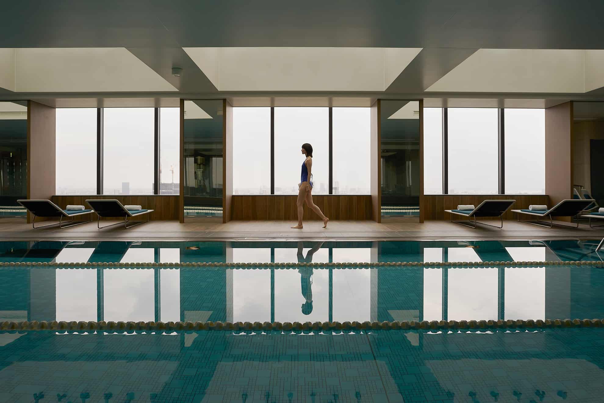 Donna di profilo che cammina accanto a piscina da nuoto coperta, oltre grandi finestre e poltrone