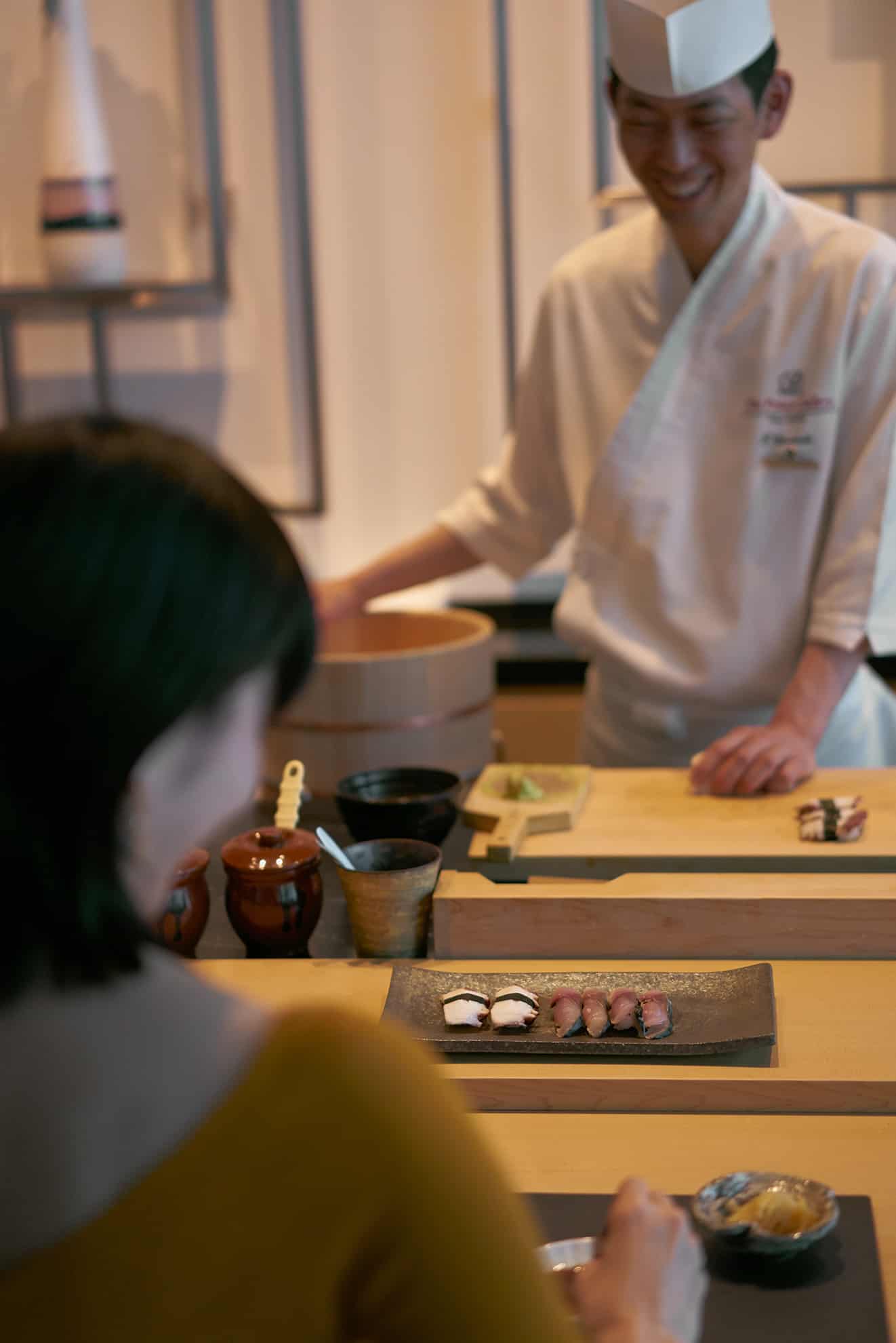Over-the-shoulder shot of plate of sushi in front of woman in yellow shirt and chef