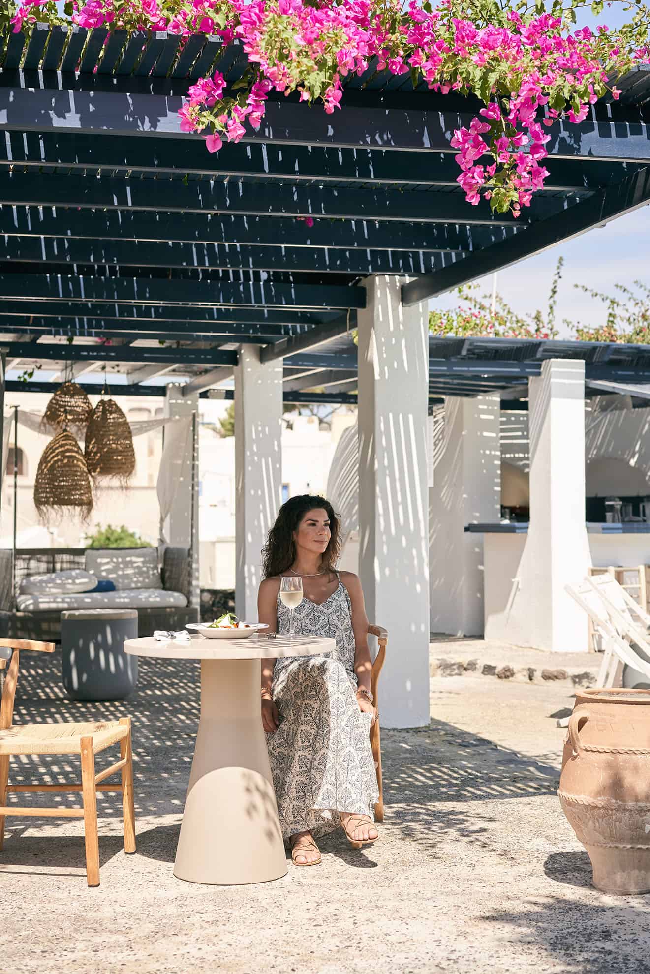 Woman in long summer dress sitting at table under a flowered pergola with a glass of wine