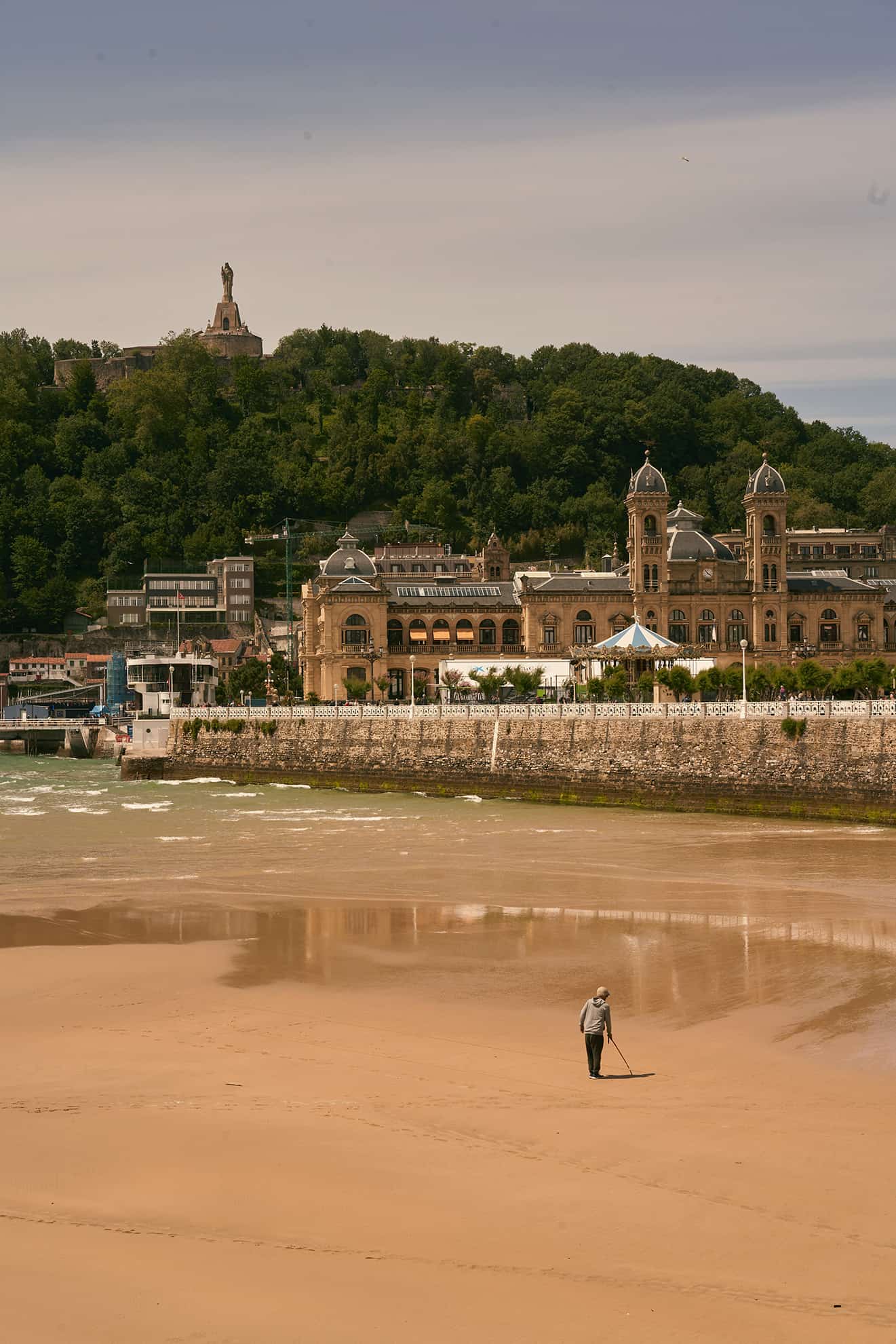 Person läuft an einem nassen Sandstrand entlang, im Hintergrund eine europäische Stadt und Wald