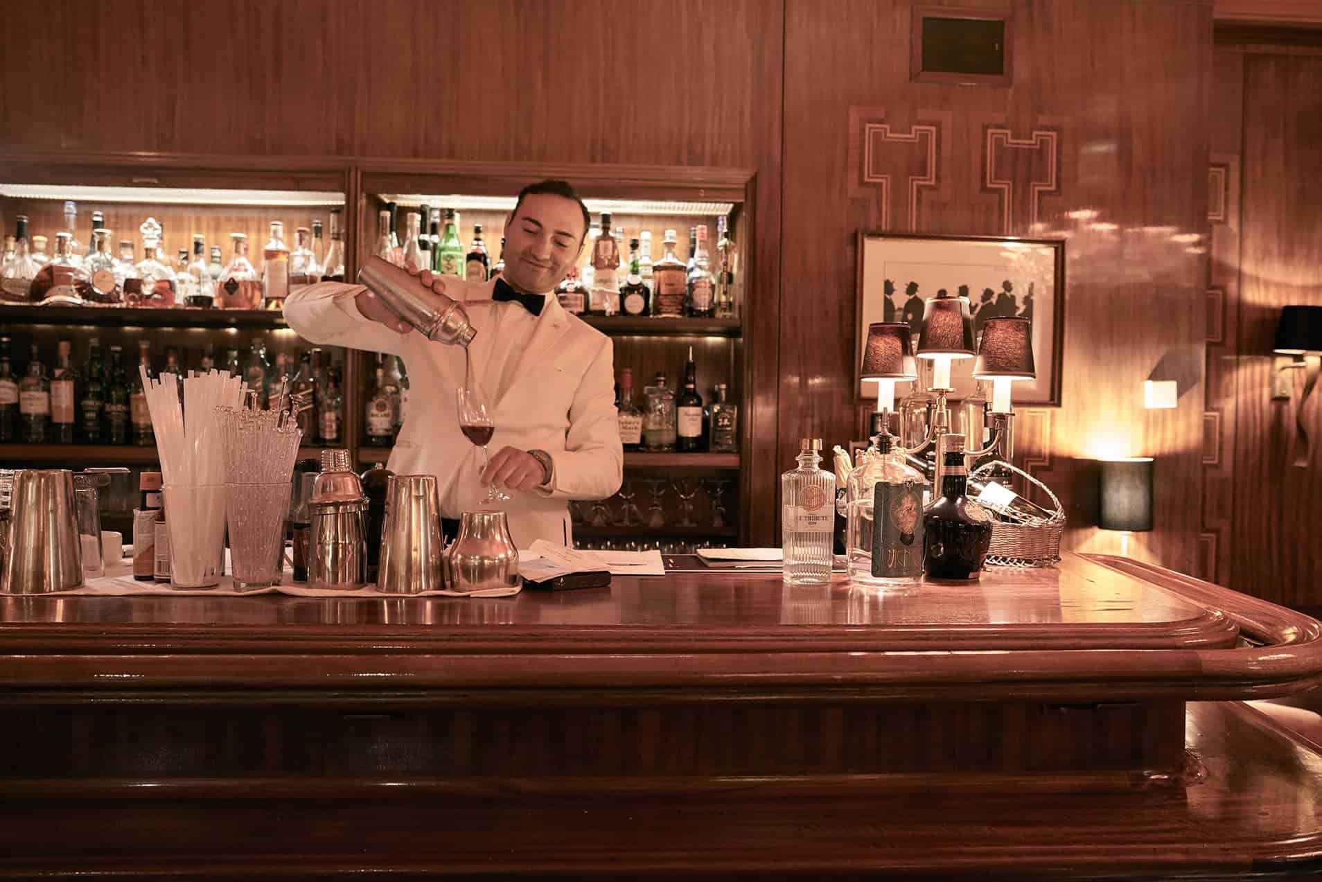 Bartender de pé atrás de um bar totalmente equipado, despejando um coquetel de uma coqueteleira de aço inoxidável