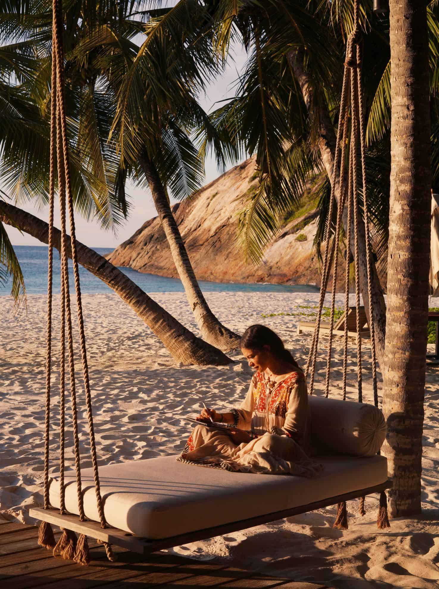 Woman writing on beach sitting on lounger hanging from palm trees