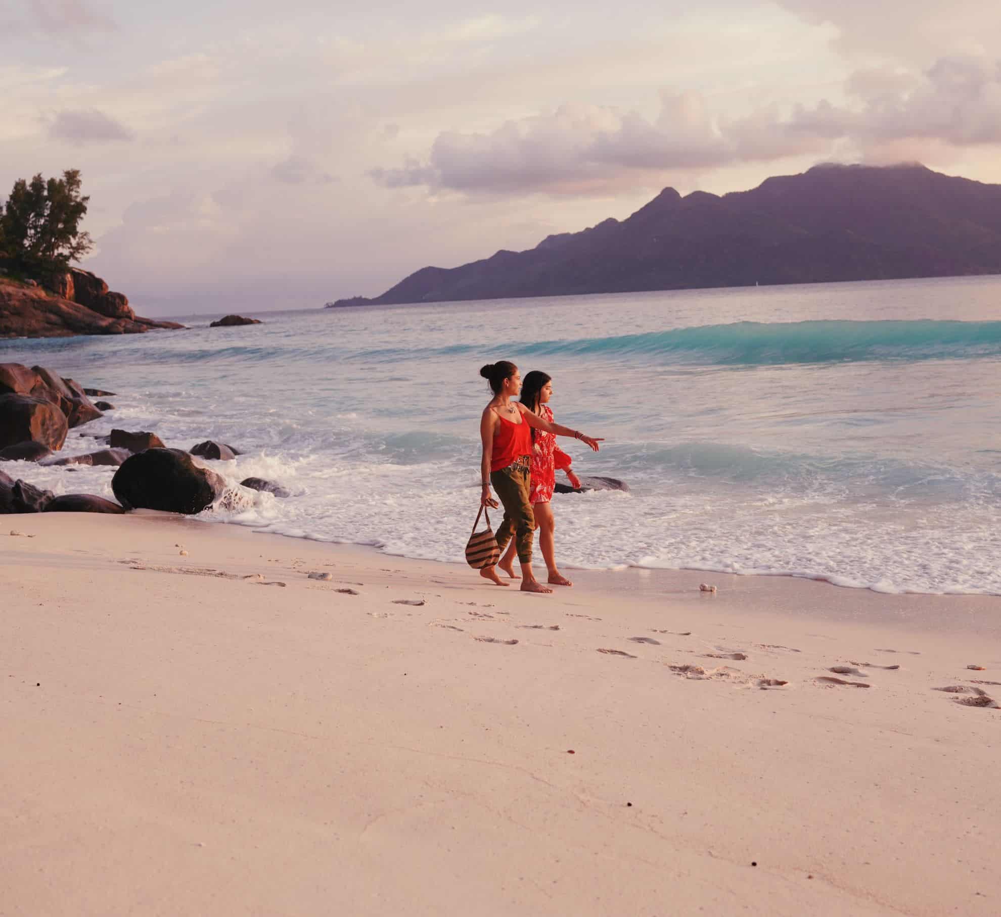 Two women walking along the beach looking out at water.