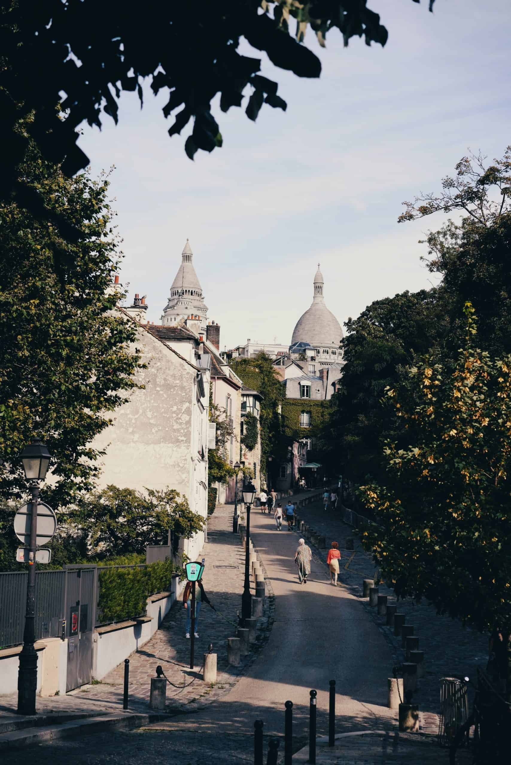 A small city street with people walking and biking; steepled buildings in the distance