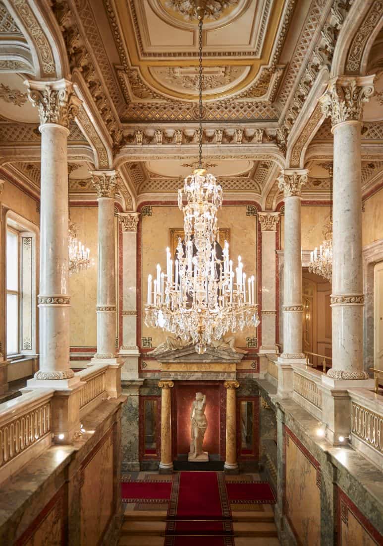 Lit crystal chandelier hanging from ornate ceiling between stone pillars with marble statue below